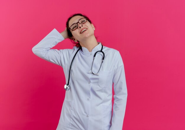 Doctora joven complacida con bata médica y estetoscopio y gafas poniendo la mano detrás de la cabeza con los ojos cerrados aislados en la pared rosa con espacio de copia