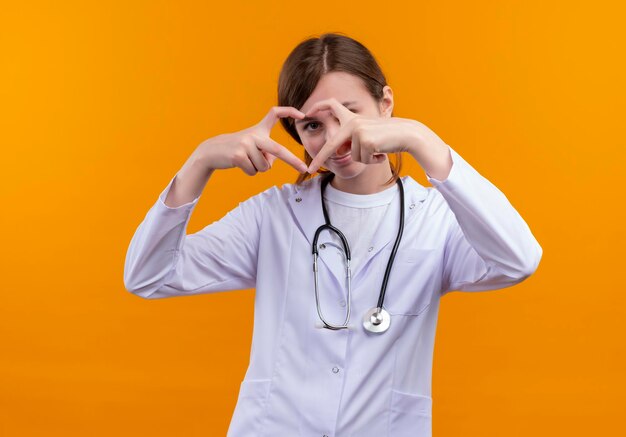 Doctora joven complacida con bata médica y un estetoscopio haciendo el signo del corazón y mirando a través de él en un espacio naranja aislado