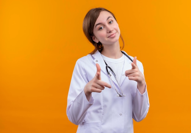 Doctora joven complacida con bata médica y un estetoscopio haciendo su gesto en un espacio naranja aislado con espacio de copia