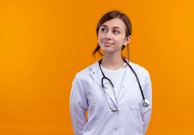 Doctora joven complacida con bata médica y un estetoscopio mirando hacia el lado izquierdo en el espacio naranja aislado con espacio de copia