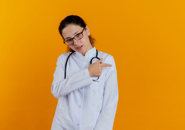 Doctora joven complacida con bata médica y estetoscopio con puntos de gafas en el lado aislado