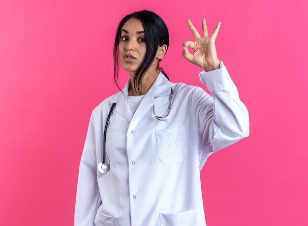 Doctora joven complacida con bata médica con estetoscopio que muestra un gesto bien aislado en la pared rosa