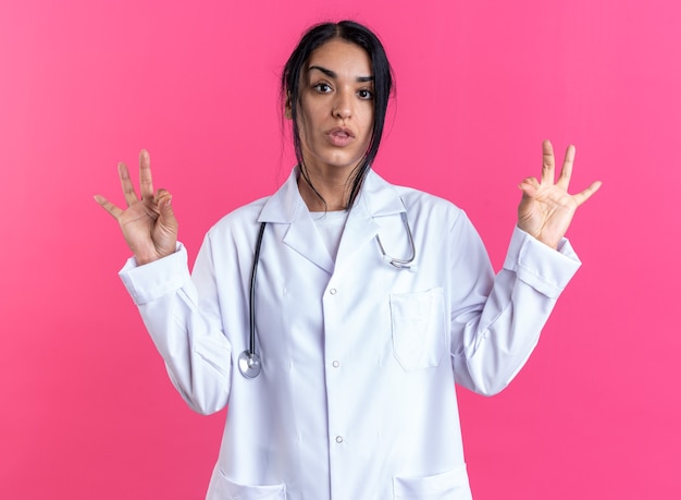 Doctora joven complacida con bata médica con estetoscopio que muestra un gesto bien aislado en la pared rosa