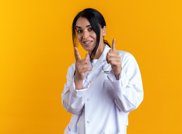 Doctora joven complacida con bata médica con puntos de estetoscopio en la cámara aislada en la pared amarilla