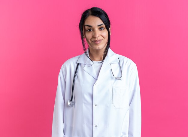 Doctora joven complacida vistiendo bata médica con estetoscopio aislado en la pared rosa