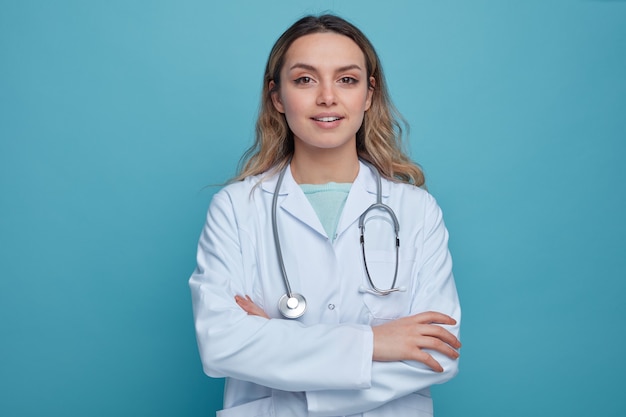 Foto gratuita doctora joven complacida vistiendo bata médica y estetoscopio alrededor del cuello de pie con postura cerrada