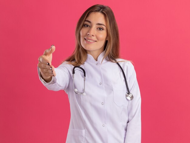 Doctora joven complacida vistiendo bata médica con estetoscopio extendiendo la mano en el frente aislado en la pared rosa