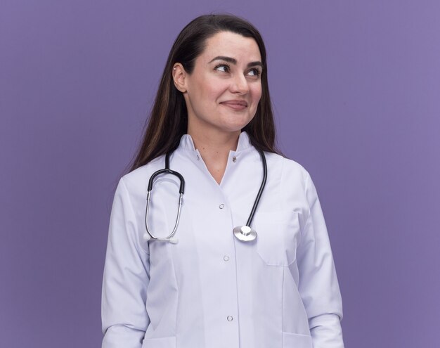 Doctora joven complacida vistiendo bata médica con estetoscopio mira al lado