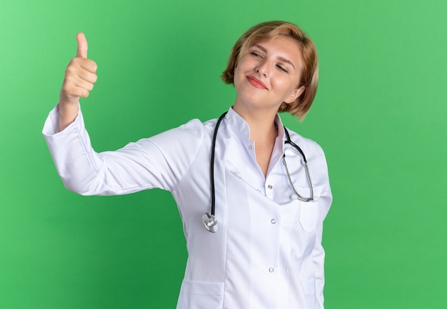 Doctora joven complacida vistiendo bata médica con estetoscopio mostrando el pulgar hacia arriba aislado en la pared verde