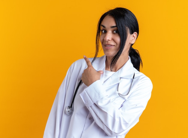 Doctora joven complacida vistiendo bata médica con puntos de estetoscopio detrás aislado en la pared amarilla con espacio de copia