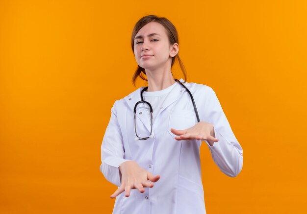 Doctora joven disgustada vistiendo bata médica y estetoscopio estirando las manos sobre la pared naranja aislada con espacio de copia