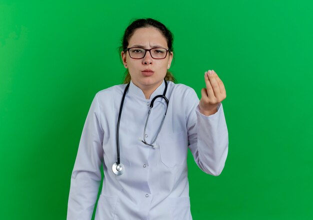 Doctora joven disgustada vistiendo bata médica y estetoscopio y gafas haciendo gesto de dinero aislado en la pared verde con espacio de copia