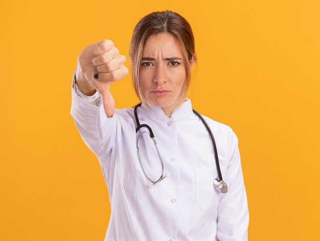 Doctora joven disgustada vistiendo bata médica con estetoscopio mostrando el pulgar hacia abajo aislado en la pared amarilla