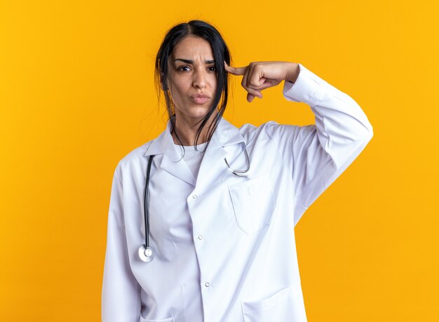 Doctora joven disgustada vistiendo bata médica con estetoscopio poniendo el dedo en la sien aislado sobre fondo amarillo