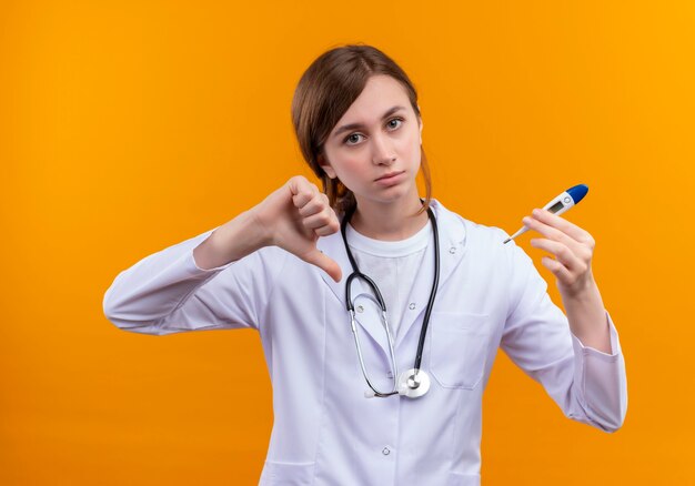 Doctora joven disgustada vistiendo bata médica y estetoscopio y sosteniendo el termómetro mostrando el pulgar hacia abajo en la pared naranja aislada