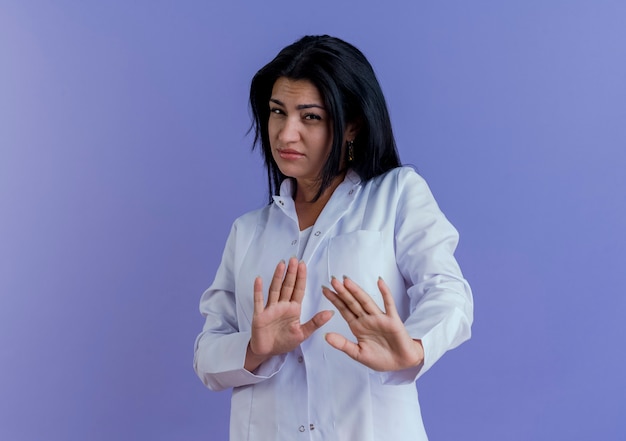 Doctora joven disgustada vistiendo bata médica sin hacer ningún gesto aislado en la pared púrpura con espacio de copia