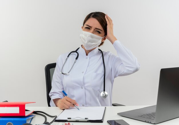 Doctora joven dolorida con bata médica y estetoscopio y máscara sentado en el escritorio con herramientas médicas y portátil poniendo la mano en la cabeza sosteniendo la pluma aislada en la pared blanca