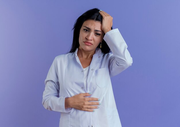 Doctora joven dolorida triste vistiendo bata médica mirando hacia abajo poniendo las manos en la cabeza y el vientre