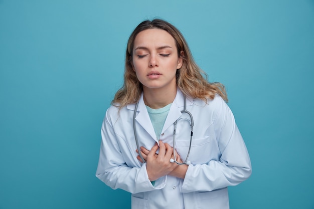 Doctora joven dolorida vistiendo bata médica y un estetoscopio alrededor del cuello manteniendo las manos en el pecho con los ojos cerrados