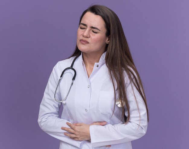 Doctora joven dolorida vistiendo bata médica con estetoscopio pone las manos sobre el vientre