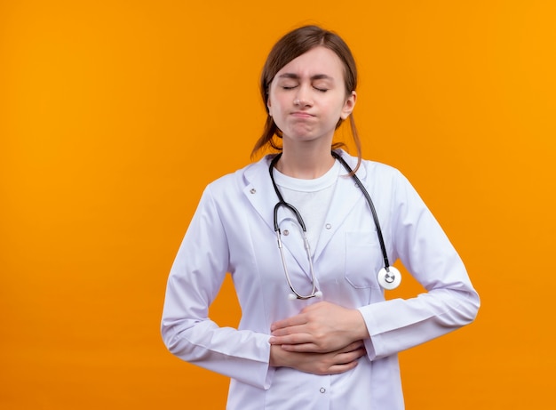 Doctora joven dolorida vistiendo bata médica y estetoscopio poniendo las manos sobre el vientre con los ojos cerrados en el espacio naranja aislado con espacio de copia