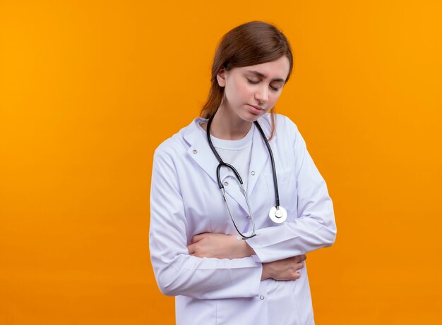 Doctora joven dolorida vistiendo bata médica y estetoscopio poniendo las manos sobre el vientre con los ojos cerrados en el espacio naranja aislado con espacio de copia