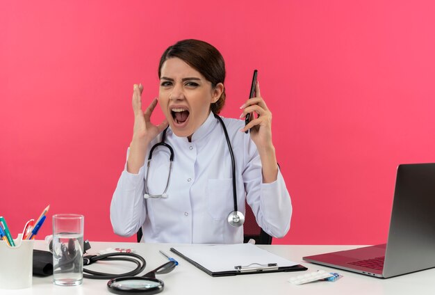 Doctora joven estresada con bata médica y estetoscopio sentado en el escritorio con herramientas médicas y computadora portátil sosteniendo el teléfono móvil manteniendo la mano en el aire y gritando aislado en la pared rosa