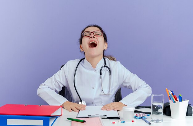 Doctora joven estresada vistiendo bata médica y estetoscopio sentado en el escritorio con herramientas médicas poniendo las manos sobre el escritorio gritando con los ojos cerrados aislados