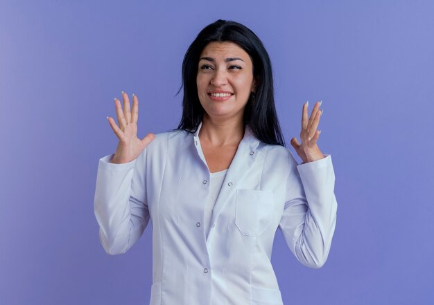 Doctora joven irritada vistiendo bata médica manteniendo las manos en el aire mirando al lado