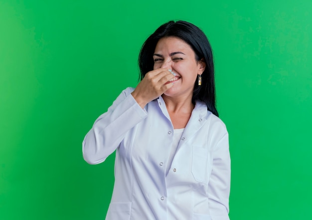 Doctora joven irritada vistiendo bata médica sosteniendo la nariz aislado en la pared verde con espacio de copia
