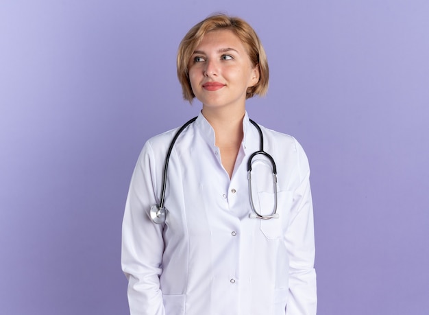 Doctora joven de lado mirando complacido vistiendo bata médica con estetoscopio aislado en la pared azul