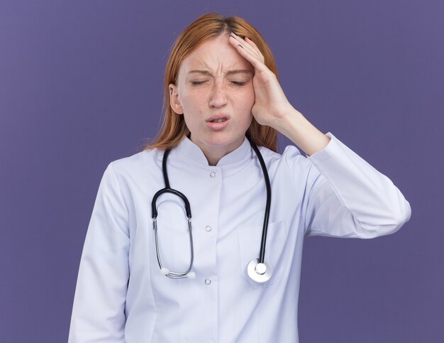 Doctora joven médico de jengibre vistiendo una bata médica y un estetoscopio poniendo la mano en la cabeza con los ojos cerrados aislados en la pared púrpura