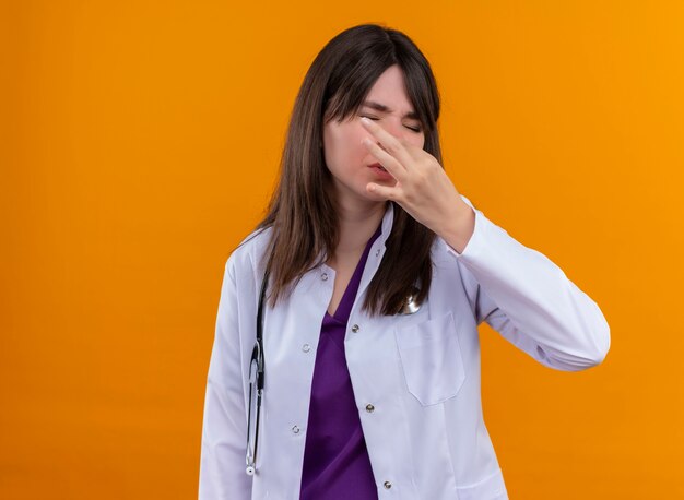 Doctora joven molesta en bata médica con estetoscopio huele gesto sobre fondo naranja aislado con espacio de copia