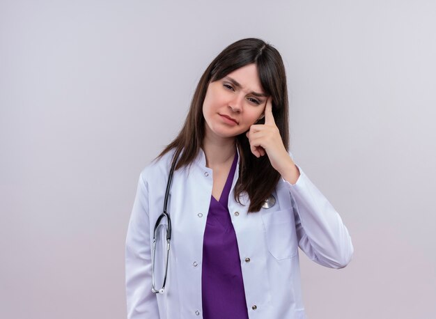 Doctora joven molesta en bata médica con estetoscopio pone el dedo en la sien sobre fondo blanco aislado con espacio de copia