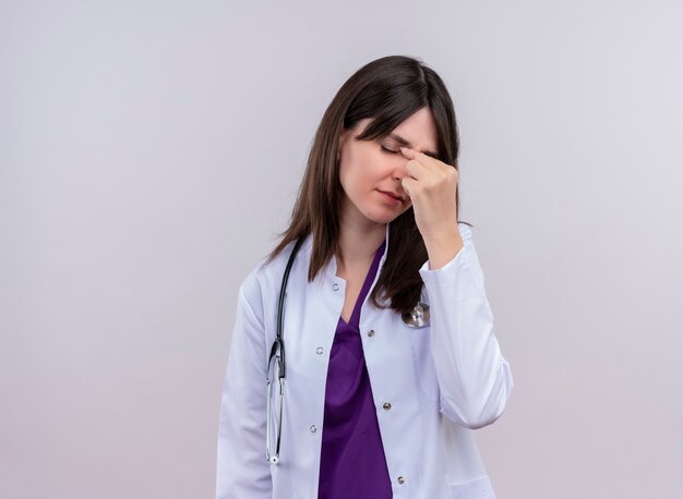 Doctora joven molesta en bata médica con estetoscopio pone la mano en la cara sobre fondo blanco aislado con espacio de copia