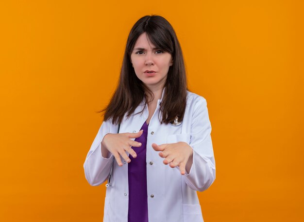 Foto gratuita doctora joven molesta en bata médica con gestos de estetoscopio para alejarse de mí sobre fondo naranja aislado con espacio de copia