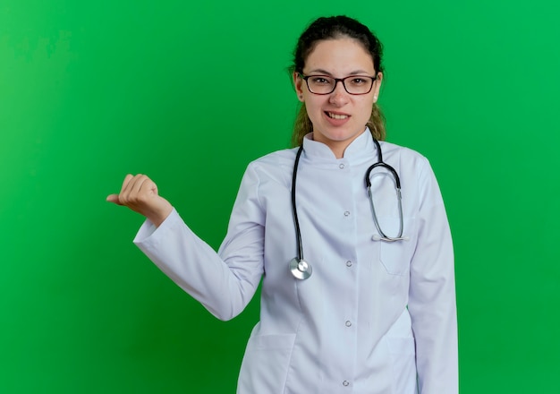 Doctora joven molesta vistiendo bata médica y estetoscopio y gafas apuntando al lado aislado en la pared verde con espacio de copia