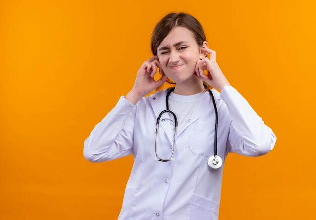 Doctora joven molesta vistiendo bata médica y estetoscopio poniendo las manos en los oídos en el espacio naranja aislado con espacio de copia