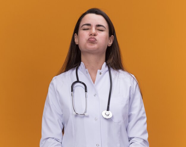 Doctora joven molesta vistiendo bata médica con estetoscopio sopla las mejillas y se para con los ojos cerrados aislados en la pared naranja con espacio de copia