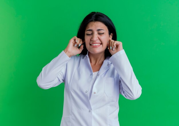 Doctora joven molesta vistiendo bata médica poniendo los dedos en los oídos con los ojos cerrados