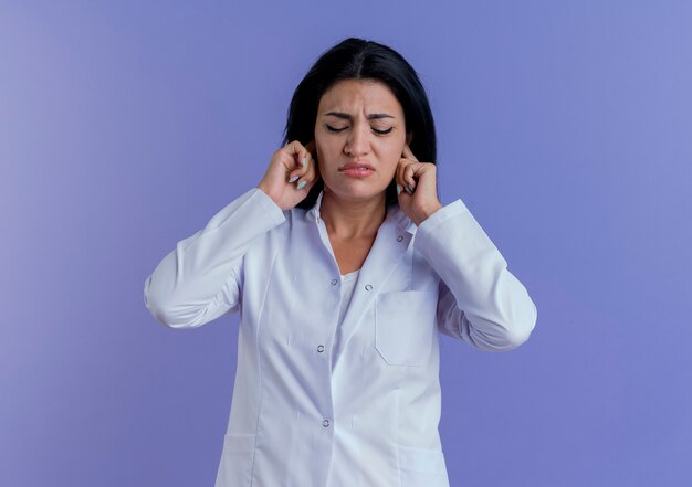 Doctora joven molesta vistiendo bata médica poniendo las manos en los oídos con los ojos cerrados