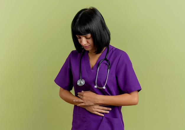 Foto gratuita doctora joven morena dolorida en uniforme con estetoscopio se encuentra con los ojos cerrados sosteniendo el vientre con las dos manos