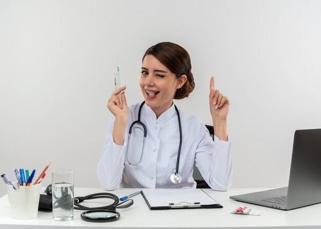 Doctora joven parpadeó con bata médica con estetoscopio sentado en el escritorio de trabajo en la computadora con herramientas médicas sosteniendo los puntos de la jeringa y mostrando la lengua en la pared blanca con espacio de copia