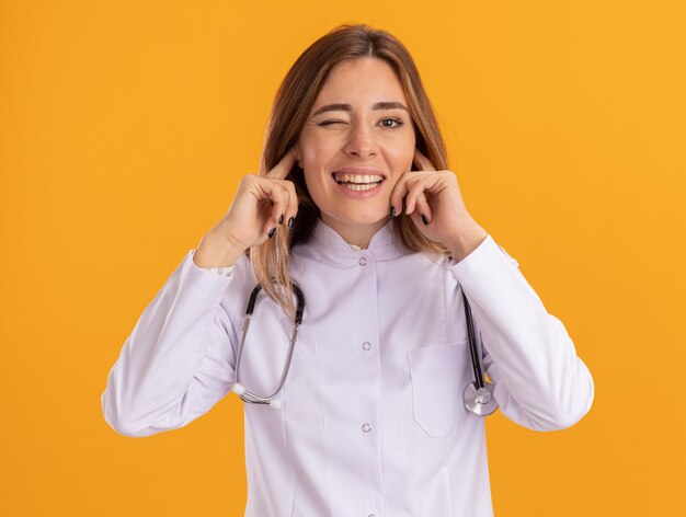 Doctora joven parpadeó vistiendo bata médica con estetoscopio orejas cerradas aisladas en la pared amarilla