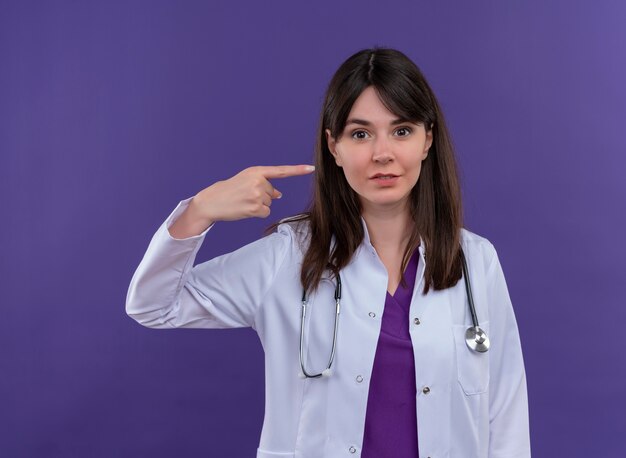 Doctora joven sorprendida en bata médica con estetoscopio apunta hacia el lado sobre fondo violeta aislado con espacio de copia
