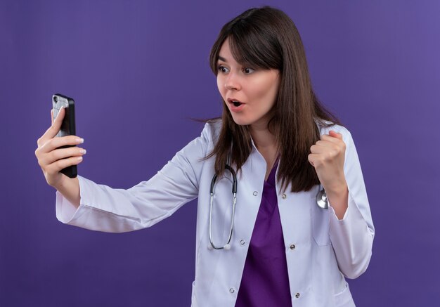 Doctora joven sorprendida en bata médica con estetoscopio mira el teléfono y mantiene el puño en alto sobre fondo violeta aislado