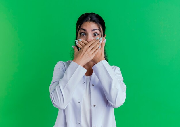 Doctora joven sorprendida vistiendo bata médica cubriendo la boca con las manos aisladas en la pared verde con espacio de copia