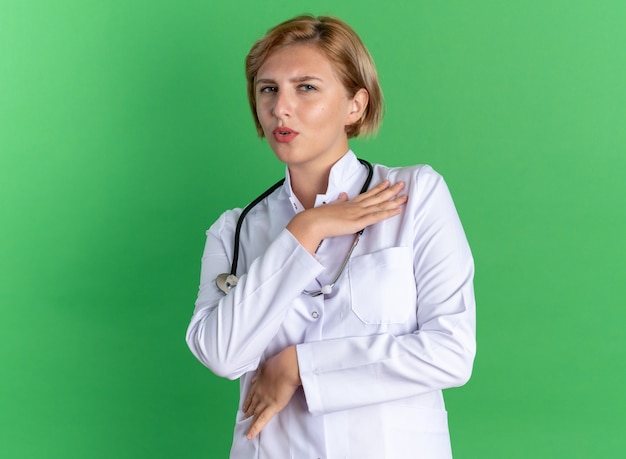 Doctora joven sorprendida vistiendo bata médica con estetoscopio aislado sobre fondo verde