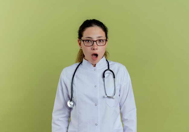 Doctora joven sorprendida vistiendo bata médica y estetoscopio con gafas aisladas en la pared verde oliva