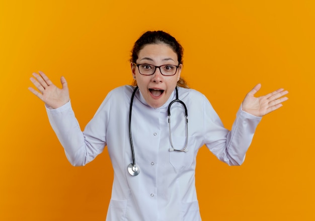 Doctora joven sorprendida vistiendo bata médica y estetoscopio con gafas extendiendo las manos aisladas en la pared naranja